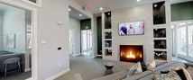 This single-floor amenity space is thoughtfully designed to serve as both a social and functional hub. The main lounge area centers around a marble-surround fireplace flanked by built-in display shelving, with a wall-mounted television above. Plush, neutral-toned sectional seating, accent pillows, and soft carpeting create a welcoming atmosphere, while a shuffleboard table and large windows providing natural light add to the relaxed yet refined ambiance. A bold green accent wall with an arched passage leads toward the entrance, adding a striking visual contrast to the light grey walls and recessed lighting throughout. Adjacent to the lounge, a glass-enclosed conference or co-working room offers a dedicated space for meetings, furnished with a long table and bar-height seating. A separate bar or social area features a dramatic textured tile accent wall, a deep navy high-top table surrounded by mixed upholstered bar stools, and eye-catching geometric pendant chandeliers that serve as a focal point. A wet bar with cabinetry and counter space completes the entertaining setup. Together, these areas form a cohesive, multi-functional community space designed for both relaxation and productivity.