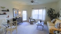 This single-floor living space offers a clean, open layout bathed in natural light from multiple large windows dressed with sheer white curtains and horizontal blinds. A ceiling fan with a light fixture is centrally positioned overhead, providing both airflow and ambient lighting throughout the room. The space is finished with wall-to-wall carpeting in a neutral tone, contributing to a warm and comfortable atmosphere. A glass-paneled door provides direct access to the outdoors, further enhancing the sense of openness and connectivity to the exterior. The room accommodates a tasteful arrangement of furnishings, including a zebra-print accent chair and a sleek glass-topped coffee table with a metal frame. A dark mirrored dresser along one wall is paired with a table lamp and a framed piece of wall art, adding a decorative focal point to the space. An additional chair with a bamboo-style frame contributes to the eclectic yet cohesive design aesthetic. The light-toned walls reflect natural light effectively, making the room feel spacious and inviting throughout the day.
