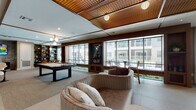 This thoughtfully designed single-floor amenity lounge offers a dynamic blend of social and leisure spaces, making it an inviting hub for gathering and recreation. The space is anchored by a striking decorative ceiling featuring warm, woven-pattern copper-toned panels accented by wooden beam detailing, lending the interior a refined yet approachable character.

The main lounge area is furnished with curved, plush seating arrangements and an array of curated accent pieces, creating a comfortable and visually engaging environment. A full-size billiards table serves as a centerpiece for recreation, complemented by a dedicated dining table with wicker-style chairs and built-in shelving stocked with books and décor. Floor-to-ceiling windows line one side of the space, flooding the interior with natural light and offering direct views of an outdoor pool and surrounding courtyard.

A built-in window bench provides additional casual seating along the glazed wall, while a cozy media nook at one end of the lounge features a large wall-mounted television set against a dramatic marble accent wall, paired with a blush sofa and sculptural accent chair.

The space also includes a well-appointed bar area with a generous white countertop, teal tile detailing, and gold-accented bar stools beneath the signature patterned ceiling. A large mirror behind the bar enhances the sense of depth and light throughout. The flooring transitions between large-format tile and plush carpet, clearly delineating each functional zone within the lounge.