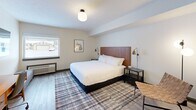 Located at 461 Munson Ave in Traverse City, Michigan, this thoughtfully designed single-floor room offers a compact yet functional layout with one bedroom and one bathroom. The main living area features light wood-style flooring, crisp white walls, and a globe ceiling fixture that creates a warm, inviting ambiance. A king-sized bed with a rich wood headboard anchors the room, flanked by matching dark nightstands with adjustable brass reading lamps. A plaid area rug adds texture and warmth underfoot. A window allows natural light to fill the space, offering a view of the surrounding area. The room also includes a mid-century modern wooden desk with a cushioned office chair, a wall-mounted flat-screen television, and a bench seat at the foot of the bed — making it well-suited for both relaxation and productivity. Sage green double doors with paneled detail provide a stylish architectural focal point, leading to a vanity and wet area. The bathroom is efficiently laid out with a walk-in glass-enclosed shower, a toilet, pedestal sink, and wall-mounted shelving for towels and toiletries, all finished in a clean, light neutral palette. A mini-refrigerator rounds out the practical amenities of this well-curated space.
