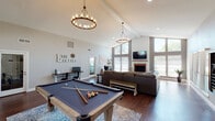 Located at 5450 Texas Ave in Abilene, Texas, The Landing is a generously sized single-floor space designed for recreation, relaxation, and gathering. The interior is defined by dramatic vaulted ceilings with exposed beams, creating an airy, open atmosphere throughout. Rich hardwood flooring runs the length of the space, complementing the neutral gray walls and white trim for a clean, modern-rustic aesthetic.

The main area serves as both a game room and lounge, anchored by a full-size pool table set atop a decorative area rug and illuminated by a pair of statement wagon-wheel chandeliers. The living area features a large sectional sofa oriented toward a shiplap-accented fireplace with a mounted flat-screen television above. Floor-to-ceiling windows and multiple sets of French doors flood the space with natural light and provide views of the outdoor surroundings.

A dedicated relaxation corner includes a massage chair, open shelving, and a workspace with a desk. French doors along one wall lead to an outdoor patio area, seamlessly connecting the interior to the exterior. A compact kitchen is visible through an open passageway, rounding out the functional offerings of the space. The overall ambiance is welcoming, versatile, and well-suited for social and recreational use.