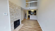 This two-story space makes an immediate impression with its dramatic double-height ceiling in the main living area, creating a sense of openness and volume that extends upward to a second-floor loft. The upper level is framed by a white metal railing, offering an open overlook to the living space below and adding architectural interest to the overall design.

The focal point of the living area is a floor-to-ceiling stacked-stone fireplace with a sleek black surround, combining a natural, textured aesthetic with a contemporary finish. Warm-toned wood-look flooring runs throughout the main level, contributing to a cohesive and inviting atmosphere.

Adjacent to the living area, the kitchen features dark espresso cabinetry paired with stainless steel and black appliances, including a range and refrigerator. A peninsula provides additional prep and casual dining space, while light-colored countertops offer contrast to the darker cabinetry.

A staircase connects the main level to the upper floor, positioned centrally and accessible from the living area. Together, the open layout, soaring ceilings, stone fireplace, and two-story design create a modern and functional living environment.