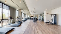 Located at 536 W Tremont Ave in Charlotte's vibrant South End neighborhood, this fitness center offers a generously proportioned, single-floor workout facility designed with both form and function in mind. The space is defined by its striking floor-to-ceiling windows that line the perimeter walls, flooding the interior with abundant natural light and offering open views of the surrounding neighborhood. Exposed concrete columns add an industrial character that complements the warm hardwood-style flooring throughout.

The cardio zone is well-equipped with a lineup of TRUE treadmills, ellipticals, stationary bikes, stair climbers, and rowing machines, thoughtfully arranged to maximize space and airflow. Ceiling fans and recessed lighting maintain a comfortable atmosphere during workouts. The strength training area features cable machines, a multi-function rack, benches, and a well-stocked dumbbell station, all reflected in large mirrored walls that visually expand the space and allow for form checks during exercise.

A dedicated accessories section houses resistance bands, medicine balls, and other functional training tools on organized shelving. The overall design aesthetic is modern and clean, with pendant Edison-style bulbs adding a warm, upscale ambiance. The open layout and generous scale of the space create an inviting, professional-grade fitness environment.
