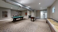Located at 649 Alden St in Fall River, Massachusetts, this single-floor amenity space is designed for entertainment and social activity, offering a well-appointed combination of a game room and a private screening room.

The main game room is a generously sized open area finished with patterned carpet tiles throughout. It features exposed industrial-style ductwork running along the ceiling, recessed lighting, and neutral gray walls accented by framed movie posters. The space accommodates a full-size pool table with overhead pendant lighting, a foosball table, and a shuffleboard table, making it a versatile hub for recreation. A wall-mounted flat-screen television is also present, along with high-top tables and bar-height seating positioned along the perimeter.

Adjacent to the game room is a dedicated home theater space furnished with rows of plush leather recliner chairs, providing a true cinema-style experience. The room features recessed lighting, sconce wall fixtures, and small transom windows that allow in subtle natural light. Exposed ductwork continues the industrial design aesthetic into this space as well.

The overall ambiance is modern and polished, with a cohesive gray-toned color palette and clean white baseboards unifying both areas. Glass-paneled entry doors provide a refined entryway into the space, completing the upscale amenity feel.