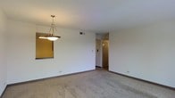 This single-floor space presents a clean, minimalist interior characterized by neutral tones and practical design. The main living area is finished with wall-to-wall carpeting in a warm beige hue, providing a comfortable underfoot feel throughout. White walls and a smooth white ceiling create a bright, open atmosphere, while dark brown baseboards add a subtle contrast that grounds the room visually.

A pendant light fixture with a bowl-shaped frosted glass shade hangs from the ceiling, serving as a central lighting element. A mirrored panel is mounted on one wall near the dining area, adding depth and reflecting light to enhance the sense of space.

A ceiling vent indicates a central HVAC system for climate control. Toward the back of the space, a hallway leads to additional areas, with a louvered bi-fold closet door providing practical storage access. The light switch panel is conveniently positioned along the wall near the hallway entry.

Overall, the space offers a straightforward, functional layout with consistent finishes throughout, making it a versatile and easy-to-maintain environment suitable for a variety of uses.