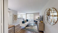 This single-floor residence offers a thoughtfully arranged layout that balances comfort and functionality across its main living areas. The living room serves as the central gathering space, featuring warm hardwood-style flooring, neutral walls, and a sliding glass door flanked by vertical blinds that floods the room with natural light and provides access to an outdoor area. A decorative macramé wall hanging and a round gold-framed mirror add character, while the open sightline to the kitchen creates a connected, airy feel.

The kitchen is equipped with white shaker-style cabinetry, dark granite countertops, and black appliances including a range and refrigerator. A pass-through counter connects the kitchen to the adjacent dining area, which features hardwood-style flooring, a large window with white blinds and sheer curtains, and an elegant brass and black chandelier overhead. A round marble-top dining table anchors the space.

The bedroom is a calm retreat with plush carpet, a distinctive teal accent wall, a window with a built-in bench seat, and ample room for furnishings. The full bathroom is conveniently accessible from the hallway and includes a shower-tub combination with a curtain rod, toilet, and hardwood-style flooring, maintaining a clean and practical aesthetic throughout.