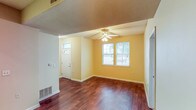 This single-story, one-bedroom, one-bathroom unit offers a functional and comfortable layout designed for efficient living. The main living area features rich, dark hardwood-style flooring that extends throughout, complemented by warm yellow-toned walls and white baseboards that create a bright, inviting atmosphere. A ceiling fan with integrated lighting is centrally mounted, and a front entry door with a decorative arched transom window allows additional natural light into the space. Two windows along the exterior wall frame views of surrounding greenery, further enhancing the open feel of the living area.

The kitchen is equipped with light oak cabinetry offering ample upper and lower storage, neutral countertops, and a white electric coil-burner range with an overhead ventilation hood. The layout provides functional counter space for meal preparation. The bedroom features plush carpeting, a window overlooking the outdoors, and a textured ceiling, offering a quiet, comfortable retreat.

The bathroom is generously sized, with light tile flooring, a wood-toned vanity with granite-style countertop, and a notably practical feature: an in-unit laundry area tucked behind bifold doors, providing added convenience. Overall, the unit maintains a clean, well-maintained aesthetic throughout.