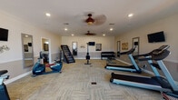 Located at 1716 Manning Dr in Clarksville, TN, this single-floor fitness center offers a well-appointed exercise space designed to support a comprehensive workout routine. The room features patterned carpet flooring throughout and is finished with crown molding and neutral-toned walls, lending a clean, polished aesthetic. Recessed lighting and two ceiling fans ensure the space remains well-lit and comfortable during use.

The main workout area is generously sized, accommodating a variety of cardio equipment including treadmills, an elliptical, a stair climber, a rowing machine, and both upright and recumbent stationary bikes. A dedicated strength training zone features a full dumbbell rack with a wide range of weights, accompanied by a wall-mounted TV and a framed mirror for form monitoring. Additional wall-mounted televisions are positioned throughout the room, allowing viewing from multiple exercise stations.

A separate lounge or stretching area at one end of the space benefits from large windows that invite natural light and offer views of the surrounding neighborhood. This section includes seating and decorative touches, providing a relaxed atmosphere for warm-ups or cooldowns. A dedicated door provides direct exterior access, adding convenience to the overall layout.