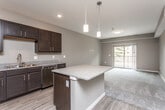 This single-floor residence is designed with a modern, open-concept layout that seamlessly connects the kitchen and living areas. The kitchen is a standout feature, appointed with rich dark-espresso cabinetry, granite countertops, and a full suite of stainless steel appliances including a refrigerator, range, microwave, and dishwasher. A central island with a matching granite surface provides additional workspace and serves as a natural dividing point between the kitchen and living space. Pendant lighting above the island adds a contemporary touch, while recessed ceiling lights provide consistent illumination throughout. Durable wood-style flooring runs through the kitchen and transitions to plush carpet in the living and bedroom areas. The living space benefits from natural light and direct access to a private balcony. The bedroom is generously proportioned, featuring a ceiling fan, a window with outdoor views, soft carpeting, and a walk-in closet with built-in shelving and rod storage. The bathroom includes a dark-cabinet vanity with granite countertop, a tub/shower combination, and wood-style flooring, maintaining a cohesive design palette throughout the unit.