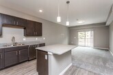 This single-floor residence is designed with a modern, open-concept layout that seamlessly connects the kitchen and living areas. The kitchen is a standout feature, appointed with rich dark-espresso cabinetry, granite countertops, and a full suite of stainless steel appliances including a refrigerator, range, microwave, and dishwasher. A central island with a matching granite surface provides additional workspace and serves as a natural dividing point between the kitchen and living space. Pendant lighting above the island adds a contemporary touch, while recessed ceiling lights provide consistent illumination throughout. Durable wood-style flooring runs through the kitchen and transitions to plush carpet in the living and bedroom areas. The living space benefits from natural light and direct access to a private balcony. The bedroom is generously proportioned, featuring a ceiling fan, a window with outdoor views, soft carpeting, and a walk-in closet with built-in shelving and rod storage. The bathroom includes a dark-cabinet vanity with granite countertop, a tub/shower combination, and wood-style flooring, maintaining a cohesive design palette throughout the unit.