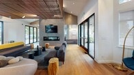 Located at 9071 E Mississippi Ave in Denver, Colorado, this single-story community clubhouse is a thoughtfully designed gathering space that blends contemporary architecture with warm, natural materials. The main lounge area serves as the heart of the space, anchored by a sleek linear fireplace set against a textured brick accent wall, with a wall-mounted television above. Soaring ceilings adorned with striking wood slat detailing add warmth and visual depth, while light hardwood flooring grounds the open-plan layout. Plush, modern seating arrangements — including curved sofas, gray sectionals, and natural wood stump accent tables — create an inviting atmosphere for socializing or relaxing. Expansive floor-to-ceiling glass doors and windows flood the interior with natural light and provide direct access to an outdoor deck furnished for al fresco gatherings. A secondary lounge area features patterned carpeting, neutral upholstered seating, and vibrant accent chairs, offering a quieter setting. A casual café-style nook with high-top tables and leather counter chairs provides a flexible workspace or social spot. The full kitchen is outfitted with light wood cabinetry, stainless steel appliances, a spacious waterfall-edge island with bar seating, and a sliding glass door leading to the outdoor area — making it well-equipped for community events and entertaining.