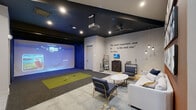 Located in Conroe, TX, this dedicated golf simulator lounge offers a well-designed space that blends sport and leisure in equal measure. The room is anchored by a full-swing golf simulator complete with a projected large-format screen, overhead projector, and a synthetic turf hitting mat, allowing users to play virtual courses with realistic ball-tracking technology. The dark ceiling contrasts sharply with the light gray walls, lending a modern, focused atmosphere ideal for play and entertainment. A Ben Hogan quote — "The most important shot in golf is the next one." — is displayed prominently on the wall, reinforcing the golf-centric character of the space. The lounge area features a comfortable gray sofa, accent chairs with gold-tone frames, and a round marble-top coffee table arranged on layered carpeting with a bold geometric pattern. Golf club racks, a media cabinet with a mounted TV, and pendant lighting add functional and aesthetic detail. An entry nook with a bistro-style table and two navy chairs sits beneath a wall-mounted display framed by striking geometric color-blocked wall art in terracotta and gray. Large panels of close-up golf ball photography serve as dramatic wall art, tying the entire theme together cohesively.