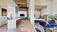 This well-appointed single-floor amenity space offers a thoughtfully designed layout that seamlessly blends social, entertainment, and professional functionality. The open main lounge is anchored by a striking accent wall with a wall-mounted TV, herringbone-patterned tile flooring, and a sleek bar counter lined with vibrant orange seating. Adjacent to the lounge, a fully equipped kitchen features warm walnut cabinetry, marble-toned countertops, stainless steel appliances including a French door refrigerator, a built-in coffee station, and a glossy tile backsplash that extends floor to ceiling. A sculptural fireplace clad in textured white tile serves as a dramatic focal point near the entrance, adding warmth and visual interest. The screening room is an immersive retreat, complete with a ceiling-mounted projector, large projection screen, deep blue velvet sofas, oversized bean bag seating, and cinematic film reel wall décor accented by ambient blue lighting. A dedicated business center offers a collaborative conference table, built-in workstations with walnut cabinetry, and wide horizontal windows that flood the room with natural light and outdoor views. Throughout, bold artwork, refined furnishings, and cohesive modern design elements create an inviting and vibrant atmosphere.