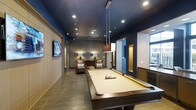 This single-floor amenity space is designed with a refined, contemporary aesthetic that balances entertainment and comfort. The centerpiece of the room is a full-size billiards table, illuminated by sleek pendant lighting that draws the eye and anchors the space. Deep navy blue walls and ceilings create a dramatic, immersive atmosphere, complemented by warm ambient lighting from stylish floor lamps positioned throughout. Large-format tile flooring in a neutral tone grounds the space with a sense of solidity and polish. A pair of rich leather armchairs are arranged invitingly near the far end of the room, offering a comfortable retreat for conversation or spectating. A wall-mounted flat-screen television adds a multimedia dimension to the lounge experience. Natural light filters in through windows along one side, softening the moody interior palette. Wood-paneled wall accents and warm-toned millwork introduce organic texture, balancing the boldness of the dark color scheme. Additional built-in cabinetry and what appears to be a beverage station are tucked to one side, enhancing the space's function as a full-service entertainment hub. Recessed ceiling lighting ensures the entire area is well-lit and versatile for various uses and gatherings.