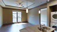 This single-floor residence showcases an open-concept design that seamlessly connects the kitchen and living areas. The living space features hardwood-style flooring that extends throughout, complemented by warm, neutral-toned walls that create a welcoming atmosphere. A tray ceiling adds architectural dimension overhead, while a ceiling fan with a light fixture provides both style and functionality.

Natural light flows generously into the living area through large windows, offering views of the surrounding exterior. The windows feature dark framing that provides a clean contrast against the lighter walls.

The kitchen area is defined by elegant granite countertops with rich, varied toning. A stainless steel sink is integrated into the countertop, and pendant lights hang above, matching the lighting style carried throughout the space. These cylindrical pendant fixtures provide focused illumination while contributing to the overall modern aesthetic.

The open layout between the kitchen and living area enhances the sense of spaciousness, making the residence feel airy and well-proportioned. The consistent use of quality finishes, including the granite surfaces and hardwood-style flooring, gives the space a cohesive and refined character.