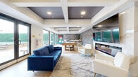 Located at 20 Grand Ave in Englewood, NJ, this contemporary lounge space offers a thoughtfully designed single-floor layout that blends comfort and modern aesthetics seamlessly. The living area is anchored by a bold navy blue tufted sofa and crisp white accent chairs with gold-tipped legs, all arranged atop a marbled area rug that adds visual texture to the light hardwood flooring beneath. A striking electric fireplace set into a white marble surround serves as a focal point, topped by a wall-mounted television and flanked by dramatic vertical stone-patterned wall panels in tones of grey and taupe. The ceiling features a distinctive coffered design with dark recessed panels and recessed lighting, adding architectural depth to the space. A chevron-patterned accent wall frames the wet bar and billiards table area toward the rear, creating a defined entertainment zone. Large floor-to-ceiling windows and glass double doors flood the interior with natural light and offer expansive views of the outdoor courtyard, which features lounge chairs, a covered spa, and lush greenery. A console table near the entry, decorated with coastal artwork and curated accessories, completes the welcoming atmosphere of this polished and versatile space.