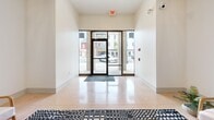 Located at 113 N Ohio Ave in Sidney, Ohio, this building lobby makes a strong first impression with its refined blend of classic and contemporary design. The main entry features floor-to-ceiling glass panels that flood the space with natural light and offer views of the surrounding streetscape, all set atop polished terrazzo flooring that flows seamlessly throughout.

The main lobby area is anchored by a bold black-and-white geometric area rug, beneath which sits a curated seating arrangement of wooden-framed accent chairs and a round side table — ideal for welcoming guests. Lush potted greenery adds warmth and a touch of nature to the environment.

Overhead, an ornate coffered ceiling with decorative pressed-tin-style panels serves as a standout architectural feature, complemented by elegant pendant and chandelier lighting fixtures. A graceful arched passageway with accent lighting leads deeper into the building, framed by distinctive teal-painted wainscoting and panel millwork that lends a sophisticated, boutique character to the corridor.

The lobby also provides convenient elevator access, housed within a stainless steel enclosure set against the teal paneled wall. A large abstract canvas painting adds an artistic focal point, tying the design elements together. This thoughtfully appointed lobby combines historic charm with modern amenities to create a polished and inviting entry experience.