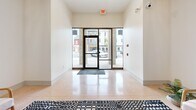 Located at 113 N Ohio Ave in Sidney, Ohio, this building lobby makes a strong first impression with its refined blend of classic and contemporary design. The main entry features floor-to-ceiling glass panels that flood the space with natural light and offer views of the surrounding streetscape, all set atop polished terrazzo flooring that flows seamlessly throughout.

The main lobby area is anchored by a bold black-and-white geometric area rug, beneath which sits a curated seating arrangement of wooden-framed accent chairs and a round side table — ideal for welcoming guests. Lush potted greenery adds warmth and a touch of nature to the environment.

Overhead, an ornate coffered ceiling with decorative pressed-tin-style panels serves as a standout architectural feature, complemented by elegant pendant and chandelier lighting fixtures. A graceful arched passageway with accent lighting leads deeper into the building, framed by distinctive teal-painted wainscoting and panel millwork that lends a sophisticated, boutique character to the corridor.

The lobby also provides convenient elevator access, housed within a stainless steel enclosure set against the teal paneled wall. A large abstract canvas painting adds an artistic focal point, tying the design elements together. This thoughtfully appointed lobby combines historic charm with modern amenities to create a polished and inviting entry experience.