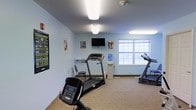 This single-floor fitness room is a dedicated exercise space designed to support cardiovascular health and overall wellness. The room features light blue walls that create a calm, energizing atmosphere, complemented by carpeted flooring that adds a layer of comfort underfoot. Overhead flush-mount lighting ensures the space is well-lit throughout.

The room is equipped with multiple pieces of cardio equipment, including treadmills and an elliptical machine, providing users with a variety of workout options. A wall-mounted flat-screen television is positioned centrally, allowing exercisers to stay entertained during their sessions. A water cooler is conveniently available to keep users hydrated, and a small desk with a chair offers a spot for quick breaks or note-taking.

Motivational fitness posters are displayed along the walls, adding an encouraging and purposeful atmosphere to the space. A large grid window along one wall allows natural light to filter in, brightening the room and providing a sense of openness. The overall layout is practical and efficient, making thoughtful use of the available floor space to accommodate a range of fitness equipment while maintaining ease of movement throughout the room.