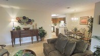 This single-story space showcases an open-concept floor plan that seamlessly connects the living room, dining area, and kitchen, creating a cohesive and functional environment. The living room is carpeted and comfortably appointed, featuring a plush sofa with decorative pillows and a wooden console table adorned with decorative wall art and greenery, adding a warm, homey touch. Adjacent to the living area, the dining space is highlighted by an elegant chandelier suspended above a round wooden dining table with ornate metal chairs, lending a classic, refined atmosphere. The kitchen, visible through an open passageway, features medium-toned wood cabinetry, white appliances, and a breakfast bar that provides additional seating and counter space. Light-toned flooring in the kitchen contrasts with the carpeted living and dining areas, helping to visually define each zone. The neutral wall tones throughout contribute to a bright, airy ambiance, while the consistent design elements unify the space. Overall, this single-floor layout offers a practical and inviting environment well-suited for everyday living and entertaining.