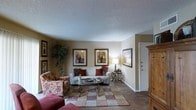 This single-story space centers around a cozy living room designed with a traditional aesthetic and a warm, neutral color palette. The room features beige walls that provide a versatile backdrop for the decor, complemented by a textured ceiling that adds subtle dimension overhead. Dark hardwood-style flooring runs throughout the space, grounded by a patterned area rug in red and cream tones that defines the seating area. The living room is furnished with a white sofa accented by red and burgundy throw pillows, paired with a floral-upholstered armchair that adds a classic, layered look. A glass-topped coffee table sits at the center, offering both style and function. Wall art is prominently displayed, with multiple framed prints arranged thoughtfully across the walls, adding visual interest and personality to the space. A tall indoor plant in the corner introduces a natural element, while a table lamp provides warm ambient lighting. A large wooden armoire near the entryway offers additional storage. The front door is conveniently positioned near the living area, making for a seamless entry into the welcoming interior.