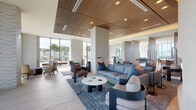 This contemporary amenity space is thoughtfully designed to offer a wide range of social and recreational experiences across a single, well-appointed floor. The main lounge area makes an immediate impression with its striking slatted wood ceiling panels, light wide-plank hardwood flooring, and floor-to-ceiling glass walls that flood the interior with natural light and frame views of the surrounding outdoor terrace and cityscape beyond.

Multiple seating zones are arranged throughout the open-plan lounge, featuring a mix of plush sofas, swivel chairs, and accent armchairs in a sophisticated palette of blues, grays, and warm taupes. A sculptural curved fireplace serves as a dramatic focal point in one section of the lounge, anchoring a comfortable conversational grouping. Textured stone wall accents complement the warm wood ceiling elements, adding depth and visual interest throughout.

A dedicated screening room offers a private cinematic experience, complete with a large projection screen, geometric LED ceiling lighting, and an array of curved lounge seating. A separate media room provides a more intimate viewing option with a wall-mounted display and dining table seating.

A sleek beverage bar, finished with marble-look countertops, a stone backsplash, and built-in cabinetry, serves as a convenient refreshment station. Carefully placed greenery and natural accents contribute to the space's refined yet welcoming ambiance.