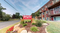 Royal Oaks Apartments offers a welcoming residential community defined by its charming exterior presentation and carefully maintained grounds. The property is introduced by a bold, iconic red entrance sign surrounded by colorful seasonal plantings, ornamental grasses, and natural boulder accents set within neatly mulched landscaping beds — creating an inviting first impression for residents and visitors alike.

The apartment building itself features a classic multi-story brick and wood-sided exterior in warm, earthy tones of red and brown. Private balconies extend from individual units, providing outdoor space with views of the surrounding neighborhood and lush tree canopy. The grounds are generously landscaped with mature oak trees and well-kept green lawns that contribute to a peaceful, park-like atmosphere throughout the community.

The setting reflects a quiet, established neighborhood environment with easy street access and a sense of suburban tranquility. The combination of durable brick construction, traditional architectural detailing, and thoughtfully designed outdoor spaces makes Royal Oaks Apartments a community that balances practicality with curb appeal. The lush greenery and seasonal flowers enhance the overall ambiance, reinforcing the community's commitment to an attractive and comfortable living environment.