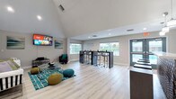 This single-floor communal space is designed with an open-concept layout that encourages social interaction and relaxation. The defining architectural feature is the dramatic vaulted ceiling, which rises to impressive heights and creates an expansive, airy atmosphere throughout the main area. Light wood-tone flooring runs seamlessly across the entire space, complementing the neutral grey and white color palette accented by vibrant teal and green décor elements.

The lounge area is anchored by a large wall-mounted flat-screen television and features comfortable white sofas, teal knit poufs, and a geometric glass coffee table atop a teal patterned area rug—ideal for gathering and entertainment. Adjacent to the lounge, a bar-height table with chrome stools provides an additional seating option.

The kitchen area is fully equipped with grey shaker-style cabinetry, white countertops, stainless steel appliances, and a decorative patterned tile island facade. Pendant lighting highlights the kitchen peninsula, which is lined with modern dark bar stools. A built-in cushioned window nook nearby adds a cozy retreat. Large windows throughout flood the space with natural light, offering views of the surrounding outdoor greenery. A separate entry with glass double doors completes the welcoming design.