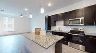Located at 6001 S Congress Ave in Austin, TX, this single-floor residence offers a thoughtfully designed open-concept layout with contemporary finishes throughout. The kitchen serves as the centerpiece of the living space, showcasing rich dark cabinetry, granite countertops, a decorative tile backsplash, and a full suite of stainless steel appliances including a refrigerator, range, and over-the-range microwave. A central island with pendant lighting provides additional counter space and a natural gathering point that flows seamlessly into the main living area. Light wood-tone luxury vinyl plank flooring runs throughout the main living spaces, complementing the clean white walls and recessed lighting that keep the interiors bright and airy. The living area benefits from a ceiling fan and natural light through large windows with blinds. The bedroom features soft carpeting, a ceiling fan, and a generous window that invites natural light. The bathroom is finished with matching granite countertops, dark wood cabinetry, a single undermount sink, a framed mirror, and a glass-enclosed walk-in shower with built-in shelving. The overall design strikes a balance between modern aesthetics and functional comfort, making this a well-appointed space in South Austin.