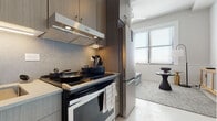 This well-appointed single-floor unit offers a cohesive modern aesthetic throughout, combining contemporary finishes with functional design. The kitchen is efficiently laid out and equipped with stainless steel appliances, including a full-size refrigerator and a range with an overhead ventilation hood. Dark wood cabinetry with a textured backsplash adds depth and warmth, while light tile flooring provides a clean, polished contrast. An entry hallway connects seamlessly to the main living spaces, maintaining an open flow through the unit. The bedroom is generously proportioned, featuring large windows dressed with horizontal blinds that invite natural light and frame city views beyond. A wall-mounted television, a velvet daybed, and thoughtfully placed lighting fixtures create a comfortable and refined atmosphere. The living area continues the contemporary theme with a wood-and-glass media console, a wall-mounted television, and an arc floor lamp that adds a sculptural touch. The bathroom is finished with mosaic tile flooring and features a glass-enclosed walk-in shower with mosaic tile walls, a sleek vanity, and clean white walls that give the space a bright, spa-like quality. Throughout, the unit presents a consistent palette of neutrals, natural wood tones, and metallic accents, resulting in a space that feels both modern and inviting.