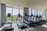 Located at 4000 Hillsboro Pike in Nashville, Tennessee, this contemporary fitness center offers a well-appointed, single-level workout facility designed to support a comprehensive exercise experience. The space is defined by dramatic floor-to-ceiling windows that flood the interior with natural light and frame stunning panoramic views of Nashville's rolling, tree-lined landscape — a standout feature that elevates the atmosphere beyond a typical gym setting. Ceiling fans and recessed lighting complement the bright, open feel throughout.

The cardio zone is equipped with a full row of treadmills, ellipticals, and stationary bikes, all positioned to take advantage of the expansive outdoor views. A dedicated strength-training area houses a wide range of resistance machines, cable systems, free weights, and stability equipment, providing options for a complete full-body workout. Structural columns with a clean, modern finish integrate seamlessly into the design without interrupting the flow of the space.

A built-in amenity station along one wall features open shelving stocked with fresh towels, closed cabinetry for storage, a compact refrigerator, and a large pass-through window overlooking the fitness floor — adding both convenience and a polished, professional aesthetic. The neutral tones, textured wall finishes, and carpeted flooring contribute to a refined, upscale ambiance throughout.