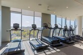 Located at 4000 Hillsboro Pike in Nashville, Tennessee, this contemporary fitness center offers a well-appointed, single-level workout facility designed to support a comprehensive exercise experience. The space is defined by dramatic floor-to-ceiling windows that flood the interior with natural light and frame stunning panoramic views of Nashville's rolling, tree-lined landscape — a standout feature that elevates the atmosphere beyond a typical gym setting. Ceiling fans and recessed lighting complement the bright, open feel throughout.

The cardio zone is equipped with a full row of treadmills, ellipticals, and stationary bikes, all positioned to take advantage of the expansive outdoor views. A dedicated strength-training area houses a wide range of resistance machines, cable systems, free weights, and stability equipment, providing options for a complete full-body workout. Structural columns with a clean, modern finish integrate seamlessly into the design without interrupting the flow of the space.

A built-in amenity station along one wall features open shelving stocked with fresh towels, closed cabinetry for storage, a compact refrigerator, and a large pass-through window overlooking the fitness floor — adding both convenience and a polished, professional aesthetic. The neutral tones, textured wall finishes, and carpeted flooring contribute to a refined, upscale ambiance throughout.