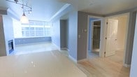 This single-floor space presents a clean, modern aesthetic defined by a thoughtful combination of materials and a neutral color palette. The main living area is finished with polished terrazzo flooring that reflects light beautifully, lending a sleek, refined character to the space. A large horizontal window along one wall allows natural light to filter in generously, brightening the blue-grey painted walls throughout.

The open layout transitions seamlessly between areas, with light hardwood flooring introduced in the adjoining section, creating a subtle yet effective visual distinction between zones. Recessed lighting and track lighting in the ceiling supplement natural light, ensuring the space remains well-lit throughout.

A central column and wall division creates a natural sense of separation between the living area and the connecting rooms. A doorway leads to what appears to be a bathroom, which features a glass-enclosed shower, suggesting a contemporary, functional design. Additional doorways hint at further rooms beyond the main living space.

White baseboards and trim provide clean contrast against the grey walls, contributing to a polished, well-finished appearance. Overall, the space conveys a calm, contemporary ambiance with practical, durable finishes throughout.