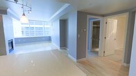 This single-floor space presents a clean, modern aesthetic defined by a thoughtful combination of materials and a neutral color palette. The main living area is finished with polished terrazzo flooring that reflects light beautifully, lending a sleek, refined character to the space. A large horizontal window along one wall allows natural light to filter in generously, brightening the blue-grey painted walls throughout.

The open layout transitions seamlessly between areas, with light hardwood flooring introduced in the adjoining section, creating a subtle yet effective visual distinction between zones. Recessed lighting and track lighting in the ceiling supplement natural light, ensuring the space remains well-lit throughout.

A central column and wall division creates a natural sense of separation between the living area and the connecting rooms. A doorway leads to what appears to be a bathroom, which features a glass-enclosed shower, suggesting a contemporary, functional design. Additional doorways hint at further rooms beyond the main living space.

White baseboards and trim provide clean contrast against the grey walls, contributing to a polished, well-finished appearance. Overall, the space conveys a calm, contemporary ambiance with practical, durable finishes throughout.