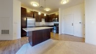 This single-floor residence offers a thoughtfully designed layout with a seamless blend of style and functionality. The kitchen serves as the centerpiece of the home, showcasing rich dark-espresso cabinetry that extends to the ceiling, light granite countertops, and a full suite of stainless steel appliances including a refrigerator, range, microwave, and dishwasher. A substantial kitchen island provides additional prep and seating space, while pendant lighting and flush-mount ceiling fixtures add warmth to the area. Warm wood-tone laminate flooring flows throughout the kitchen and connects to the adjacent living area, which transitions to plush neutral carpeting. The living space is generously sized, bathed in natural light through multiple windows fitted with horizontal blinds, and features a glass door leading to an outdoor area. The bedrooms are carpeted and well-proportioned, with one featuring a spacious walk-in closet offering built-in shelving and hanging rods. The bathroom maintains the home's consistent design aesthetic with a dark wood vanity, white countertop, framed mirror, and light hardwood-style flooring. Clean white walls and neutral finishes throughout create a bright, cohesive atmosphere.