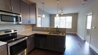This single-story space showcases a thoughtfully designed open-concept layout that seamlessly connects the kitchen and living area. The kitchen is outfitted with dark espresso-toned cabinetry, offering ample storage both above and below the countertops. Light-speckled granite countertops provide a striking contrast against the dark cabinetry, while the peninsula-style counter extends toward the living area, creating a natural divide between the two spaces.

Stainless steel appliances, including a range and dishwasher, complement the modern aesthetic. A double-basin stainless steel sink is positioned on the peninsula, offering views into the living area. Two pendant lights hang above the counter, providing focused task lighting with a contemporary touch.

The living area benefits from generous natural light streaming through large windows fitted with horizontal blinds. A ceiling fan is centrally positioned overhead, adding both functionality and a finishing detail to the space. Rich, dark hardwood-style flooring runs throughout, tying the kitchen and living areas together cohesively. The overall ambiance is modern and clean, with neutral gray walls enhancing the sense of openness.