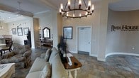 This single-floor space presents a thoughtfully designed lobby or reception area that combines classic elegance with a warm, inviting atmosphere. The room features notably high ceilings adorned with crisp crown molding, creating a sense of openness and grandeur. A striking wrought-iron chandelier with cylindrical candle-style lights serves as a central focal point, casting a warm glow throughout the space.

The flooring consists of large-format stone-look tiles in earthy, neutral tones that complement the soft beige and cream wall palette. Seating is anchored by a plush, light-toned sofa with decorative botanical-patterned throw pillows, paired with a classic turned-leg coffee table — ideal for welcoming guests comfortably.

Along one wall, a rustic wooden console table is topped with a framed ornate mirror and decorative accessories, adding a touch of traditional charm. Tall dried botanical stems provide a natural, textural accent. Wall-mounted sconce lighting further enhances the warm ambiance.

A framed informational display and a paneled white door indicate a functional transition point within the space, suggesting an office or private area beyond. The overall design balances decorative sophistication with practical functionality, making it well-suited as a welcoming reception or common gathering area.