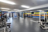 Located at 1501 Little Gloucester Rd in Blackwood, NJ, this single-floor fitness facility offers a generously sized, open workout environment designed to accommodate a broad range of exercise activities. The space is defined by its vibrant color scheme, with painted cinder block walls featuring bold horizontal stripes of blue, yellow, and orange that give the facility an energetic and motivating atmosphere.

The main workout floor is covered in durable rubber flooring, providing a safe and supportive surface for all types of training. Large wall-mounted mirrors run along a significant portion of the space, visually expanding the room and allowing users to monitor their form during exercise. Overhead fluorescent lighting ensures the entire area is well-lit throughout.

The facility is well-equipped with a diverse selection of fitness equipment. Cardio offerings include multiple treadmills and stationary bikes, while strength training needs are met with cable machines, benches, and a medicine ball rack. A dedicated area features freestanding Title-brand heavy punching bags, supporting boxing and martial arts training. Curtained sections along the walls provide additional flexible use areas. Wall-mounted televisions are positioned throughout for entertainment. The layout encourages open-floor movement while keeping equipment zones clearly defined, making the facility functional for both individual workouts and group training sessions.