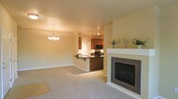 This single-level space is designed with an open-concept layout that seamlessly connects the living area, dining space, and kitchen. The living room is finished with plush wall-to-wall carpeting in a neutral beige tone and features a standout fireplace framed in dark mosaic tile with a crisp white mantel, serving as a focal point of the room. Adjacent to the living area, the dining space is illuminated by an elegant pendant chandelier that adds a refined touch to the layout.

The kitchen is partially separated by a raised peninsula with a dark countertop and seating overhang, providing both a functional workspace and casual dining area. Medium-toned wood cabinetry lines the kitchen walls, complemented by black appliances including a range and microwave. Track lighting on the kitchen ceiling ensures the workspace is well-lit. The kitchen flooring transitions to a darker surface, distinguishing it from the carpeted living and dining areas. Warm, neutral wall tones are consistent throughout, contributing to a cohesive and inviting ambiance across the entire space.