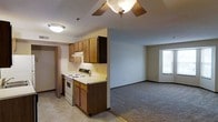 This single-floor residence offers a functional and straightforward layout that connects the kitchen and living area in an open-concept design. The kitchen is equipped with wood-toned cabinetry featuring both upper and lower storage, a white electric range with overhead ventilation, and light-colored countertops. Tile flooring in the kitchen provides a clean, easy-to-maintain surface that transitions into carpeted flooring in the adjoining living area. The living space is generously proportioned, featuring a ceiling fan with an integrated light fixture that provides both airflow and ambient lighting. Neutral-toned walls throughout lend the space a calm and understated atmosphere. A window in the living area allows natural light to fill the room, contributing to a bright and open feel. A doorway visible near the kitchen suggests additional rooms or storage areas accessible from the main living space. Overall, the layout is practical and well-organized, making efficient use of the available space while maintaining a comfortable and livable environment suitable for everyday use.