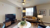 This single-floor residence offers a thoughtfully designed layout that balances comfort and practicality throughout. The living area serves as the heart of the space, featuring warm wood-look flooring, a ceiling fan, and a large window dressed with patterned curtains and blinds that invite generous natural light. The room accommodates a comfortable seating arrangement alongside a round coffee table and a wall-mounted flat-screen television, creating a welcoming and functional environment.

The kitchen is a standout feature, outfitted with crisp white shaker-style cabinetry extending to the upper walls, providing ample storage. Stainless steel appliances — including a range, over-the-range microwave, refrigerator, and dishwasher — complement the light-toned countertops. Pendant lighting in a warm yellow hue adds a modern accent, while the open pass-through connects the kitchen to the living area, enhancing the sense of openness.

The bathroom is clean and well-organized, presenting a long white vanity with a bright countertop and a large mirror. Wood-look flooring continues throughout, and a tub-shower combination with a patterned curtain completes the space. Overall, the residence presents a cohesive, modern aesthetic with warm neutral tones and practical finishes throughout.
