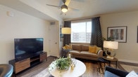 This single-floor residence offers a thoughtfully designed layout that balances comfort and practicality throughout. The living area serves as the heart of the space, featuring warm wood-look flooring, a ceiling fan, and a large window dressed with patterned curtains and blinds that invite generous natural light. The room accommodates a comfortable seating arrangement alongside a round coffee table and a wall-mounted flat-screen television, creating a welcoming and functional environment.

The kitchen is a standout feature, outfitted with crisp white shaker-style cabinetry extending to the upper walls, providing ample storage. Stainless steel appliances — including a range, over-the-range microwave, refrigerator, and dishwasher — complement the light-toned countertops. Pendant lighting in a warm yellow hue adds a modern accent, while the open pass-through connects the kitchen to the living area, enhancing the sense of openness.

The bathroom is clean and well-organized, presenting a long white vanity with a bright countertop and a large mirror. Wood-look flooring continues throughout, and a tub-shower combination with a patterned curtain completes the space. Overall, the residence presents a cohesive, modern aesthetic with warm neutral tones and practical finishes throughout.