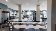 This contemporary fitness center is thoughtfully designed to deliver a complete workout experience in a spacious, single-floor layout. The space immediately stands out with its signature wave-patterned flooring in contrasting shades of navy, gray, and cream, adding a dynamic visual energy throughout the facility.

The cardio zone is lined with large floor-to-ceiling windows that flood the area with natural light and offer outdoor views, creating an open and motivating atmosphere. Equipment in this section includes treadmills, elliptical machines, rowing machines, and stationary bikes. Suspended linear pendant lighting complements the bright, airy feel of the space.

The strength training area is equally well-appointed, featuring a full dumbbell rack displayed along mirrored walls that amplify the sense of space, along with cable machines, weight benches, and multi-functional strength stations. A dedicated functional training room with glass partition doors houses TRX suspension trainers, balance equipment, and wall-mounted accessories, highlighted by a bold topographic line-art mural.

Throughout the facility, dark navy accent walls and structural columns provide architectural definition, while mounted flat-screen TVs offer entertainment during workouts. The overall ambiance is sleek, modern, and professional, making this fitness center a standout amenity.