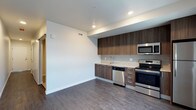 This single-floor space showcases a clean, modern design with an open layout that connects the kitchen and living areas seamlessly. The kitchen is equipped with stainless steel appliances, including a range, microwave, and dishwasher, complemented by rich, dark wood-grain cabinetry that provides ample storage. A neutral tile backsplash adds texture while maintaining a cohesive aesthetic. The countertops are finished in a light tone, creating an appealing contrast against the darker cabinetry. Recessed lighting illuminates the space efficiently, highlighting the crisp white walls and smooth ceilings. The flooring throughout features medium-toned hardwood-style planks that add warmth and continuity to the overall design. Wide baseboards provide a clean, finished look. A barn-style sliding door is visible, adding a subtle architectural detail. The open wall space adjacent to the kitchen offers flexibility for furniture arrangement, making the layout highly functional. Overall, the space presents a contemporary, well-appointed interior with a neutral palette that feels inviting and versatile.