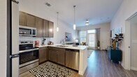 This thoughtfully designed 1-bedroom, 1-bathroom unit offers a functional and stylish open-concept layout across a single floor. The combined kitchen and living area serves as the heart of the space, featuring rich dark-brown cabinetry, a subway tile backsplash, stainless steel appliances including a range, microwave, dishwasher, and refrigerator, and a peninsula with pendant lighting overhead. The kitchen flows seamlessly into the living room, which is brightened by large windows that invite generous natural light and provide views of the outdoors. Wood-style plank flooring runs consistently throughout the unit, adding warmth and visual continuity. The bedroom is a comfortable retreat with a window allowing natural light and direct access to both the bathroom and a dedicated walk-in closet. The closet features built-in shelving and double hanging rods, offering well-organized storage. The bathroom is finished with dark cabinetry matching the kitchen, a white countertop vanity, a soaking tub with tiled surround, and a large mirror. The space presents a cohesive modern aesthetic with clean lines, neutral walls, and consistent finishes throughout, making it a practical and inviting one-bedroom residence.