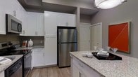 This single-floor space showcases a thoughtfully designed kitchen as its centerpiece. The kitchen features crisp white shaker-style cabinetry extending from floor to ceiling, providing generous storage throughout. Stainless steel appliances, including a full-size refrigerator, complement the modern aesthetic, while light-toned granite countertops add both functionality and visual appeal. A kitchen island with matching granite offers additional prep space and casual dining potential. The light wood-style flooring runs throughout, contributing to a warm, cohesive feel. Neutral gray walls create a versatile backdrop that ties the space together. Overhead, a contemporary flush-mount light fixture provides ample illumination. A louvered-door closet is visible beyond the kitchen, hinting at thoughtful storage solutions throughout the unit. The overall design balances practicality with modern style, featuring clean lines and a neutral color palette that creates a bright, welcoming atmosphere suited to a variety of interior design preferences.