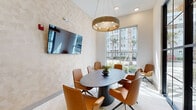 Located in Saint Johns, Florida, this single-floor quiet room facility offers a thoughtfully designed environment for focused meetings and private collaboration. The space is defined by its striking floor-to-ceiling black-framed glass partition walls, which provide both visual openness and acoustic separation while allowing natural light to flow freely throughout the interior.

The primary meeting room is centered around a sleek dark oval conference table surrounded by cognac leather chairs, creating a warm yet professional atmosphere. A large wall-mounted flat-screen display is positioned prominently for presentations and video conferencing. Overhead, a bold circular gold chandelier serves as a standout design element, adding a touch of sophistication to the otherwise clean, neutral palette.

Walls are finished with a subtle geometric textured wallcovering in soft warm tones, complementing the light-toned carpet and tile flooring. A full-height window frames exterior views of palm trees and the surrounding community, filling the room with generous Florida sunshine and a sense of openness.

The entry area features the same refined glass-and-steel framing, transitioning smoothly into a corridor that connects to additional spaces within the building. The overall ambiance is modern, polished, and purposefully calm — well-suited for productive, distraction-free gatherings.
