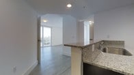 This single-floor space showcases a thoughtfully designed open-concept layout that seamlessly connects the kitchen and living areas. The kitchen features granite countertops with speckled detailing, a stainless steel sink, and a peninsula that serves as both a functional workspace and a natural divider between the cooking and living zones. Recessed lighting throughout provides consistent, even illumination across the space.

The living area benefits greatly from expansive floor-to-ceiling windows and a glass-panel balcony door, flooding the interior with natural light and offering views of the surrounding cityscape. Light-toned hardwood-style flooring runs throughout, adding warmth and continuity to the open layout.

Crisp white walls and clean architectural lines give the space a contemporary aesthetic. An arched passageway connects the main living area to additional rooms, adding a subtle architectural detail that breaks up the otherwise minimalist design. Additional doors lead to private spaces, suggesting well-defined functional zones within the unit. Overall, the space communicates a clean, modern ambiance suited for comfortable urban living.