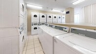Located at 8 Silver Maple Crt in Brampton, Ontario, this shared laundry and amenity facility offers a well-organized, functional space across a single floor. The laundry area is equipped with a generous row of front-loading washing machines and an array of stacked and standalone dryers, providing ample capacity for residents. Bright fluorescent lighting illuminates the space, complemented by clean, light-toned ceramic tile flooring that extends throughout. Walls are finished in a neutral warm beige, contributing to a clean and practical atmosphere. A wall-mounted television is positioned above the washers, offering entertainment while residents wait. Instructional signage and a recycling bin support ease of use and sustainability. Adjacent to the laundry area, a comfortable lounge space features a round dining table with seating, vertical blinds over windows that allow natural light to filter in, and a built-in bookshelf stocked with a wide selection of books. A digital information kiosk and a community bulletin board provide residents with convenient access to building notices and updates. A long countertop along one wall offers a practical folding or waiting surface. The facility combines utility with modest communal comfort, making it a practical and welcoming shared amenity.