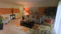 This single-floor space makes a striking first impression with its bold design choices and warm, energetic ambiance. The living area serves as the heart of the space, showcasing a distinctive color palette anchored by vivid orange accent walls that add depth and personality to the room. Soft, neutral-toned carpet flooring runs underfoot, providing a comfortable contrast to the lively wall colors.

The living room is thoughtfully arranged around a dark espresso-finished coffee table, which sits atop a large floral-patterned area rug that echoes the room's orange and green color scheme. A plush grey sofa is adorned with an assortment of colorful throw pillows in shades of orange, green, and cream, adding layers of texture and comfort. A large floral canvas painting anchors one of the accent walls, tying the room's design elements together cohesively.

Ambient lighting from a classic table lamp casts a warm glow throughout the space, enhancing its inviting character. A small entryway is visible near the front door, offering a seamless transition into the main living area. The overall ambiance is cheerful and welcoming, with a curated mix of bold color and cozy furnishings that create a lively yet comfortable environment.
